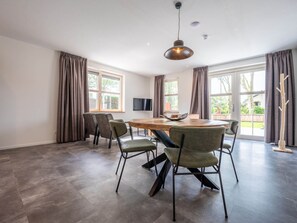 Ein geräumiges Zimmer mit einem großen runden Tisch, sechs Stühlen, einem Flachbildfernseher und großen Fenstern mit Vorhängen.