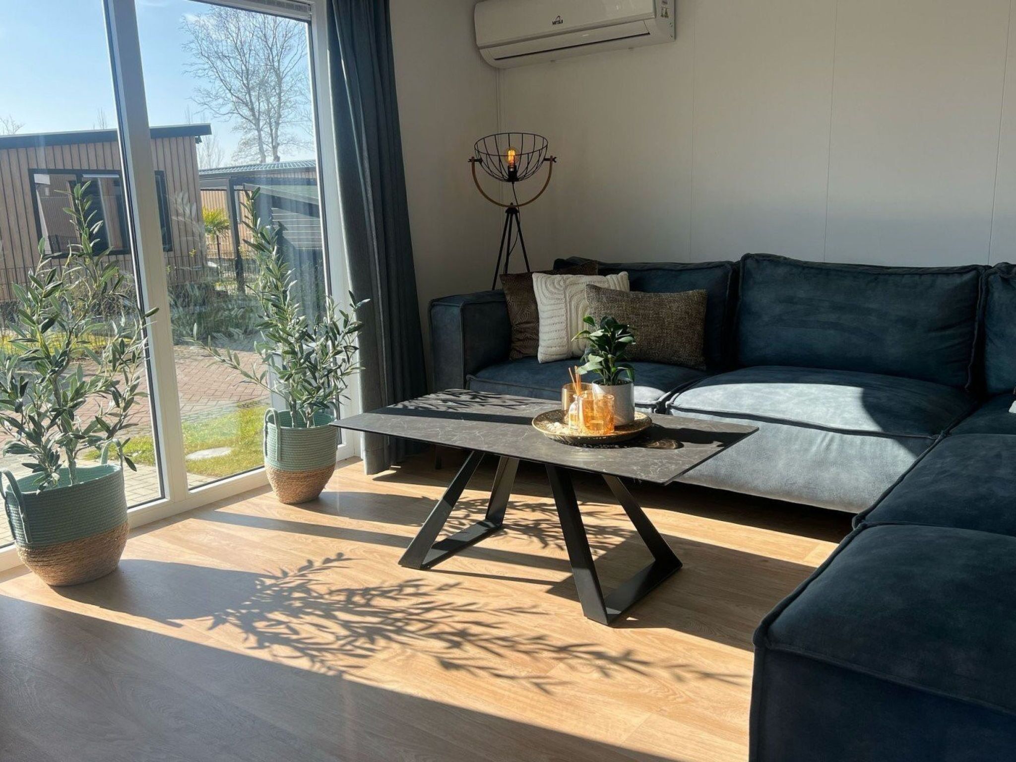 Una sala de estar moderna con un sofá, una mesa de centro y una lámpara de pie. Hay plantas en macetas y vistas al exterior.