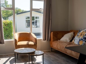 Ein Zimmer mit einer Couch, einem Sessel und einem kleinen Tisch. Es gibt Fenster mit Vorhängen und einen Blick auf ein Haus.