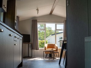 Ein Zimmer mit Küchenzeile, Fernseher, einem fensterbelaubten Fenster, einem Sessel und einem Tisch.