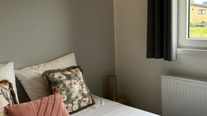 A bedroom with a bed, pillows, a radiator, and a window with curtains. - Vakantiepark de Zeeuwse Parel - Noordzee Deluxe by Interhome (Scherpenisse)