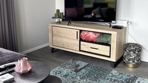 A modern living room with a flat-screen TV, a wooden cabinet with drawers, a round side table with a vase, and a patterned rug. - Vakantiepark de Zeeuwse Parel - Noordzee Deluxe by Interhome (Scherpenisse)