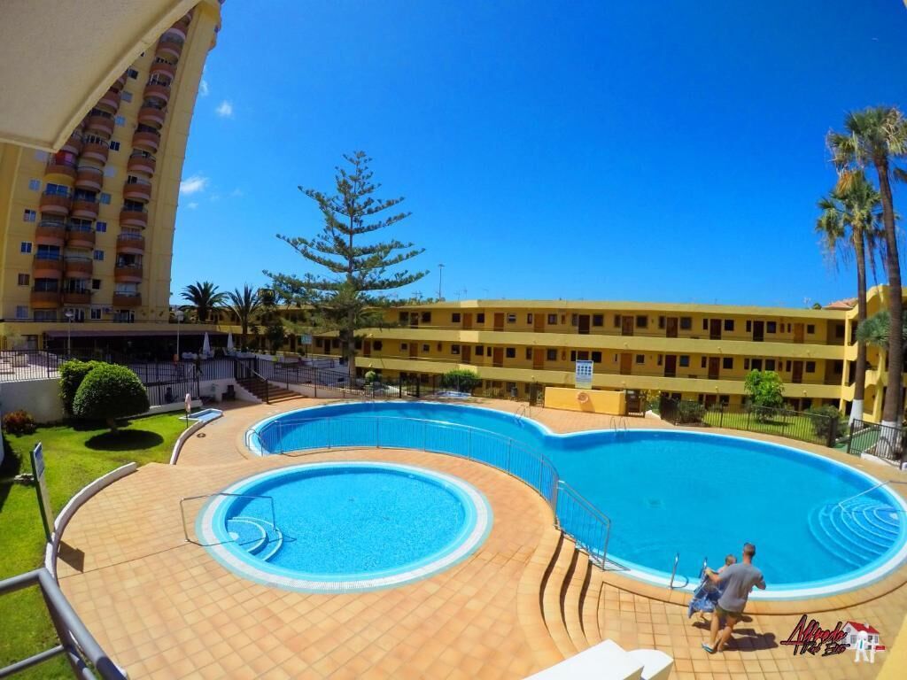 A hotel with a large swimming pool, a building with balconies, and a clear blue sky.