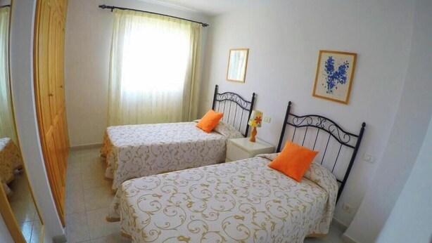 A room with two beds, each with a headboard, a bedside table with a lamp, and a window with curtains. - Torres del Sol (Arona)