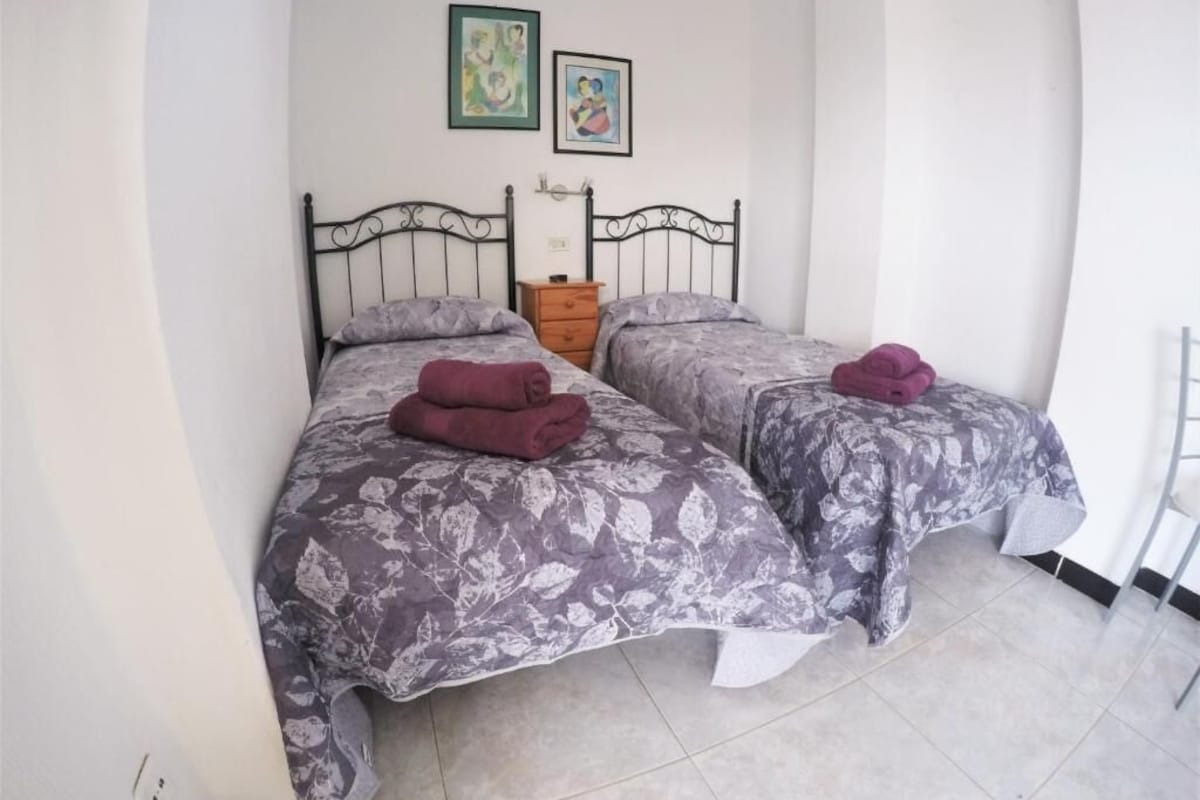 A bedroom with two beds, each with a floral-patterned bedspread and a purple towel. There are two framed pictures on the wall above the beds.