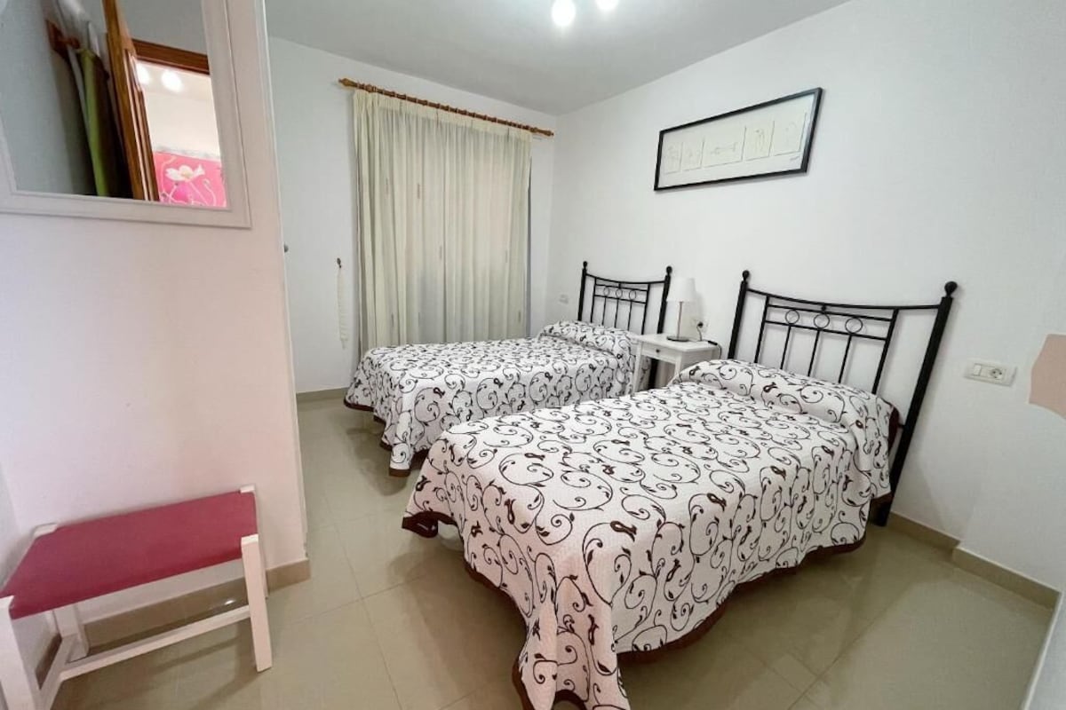 A room with two beds, a bench, a mirror, and a window with curtains.
