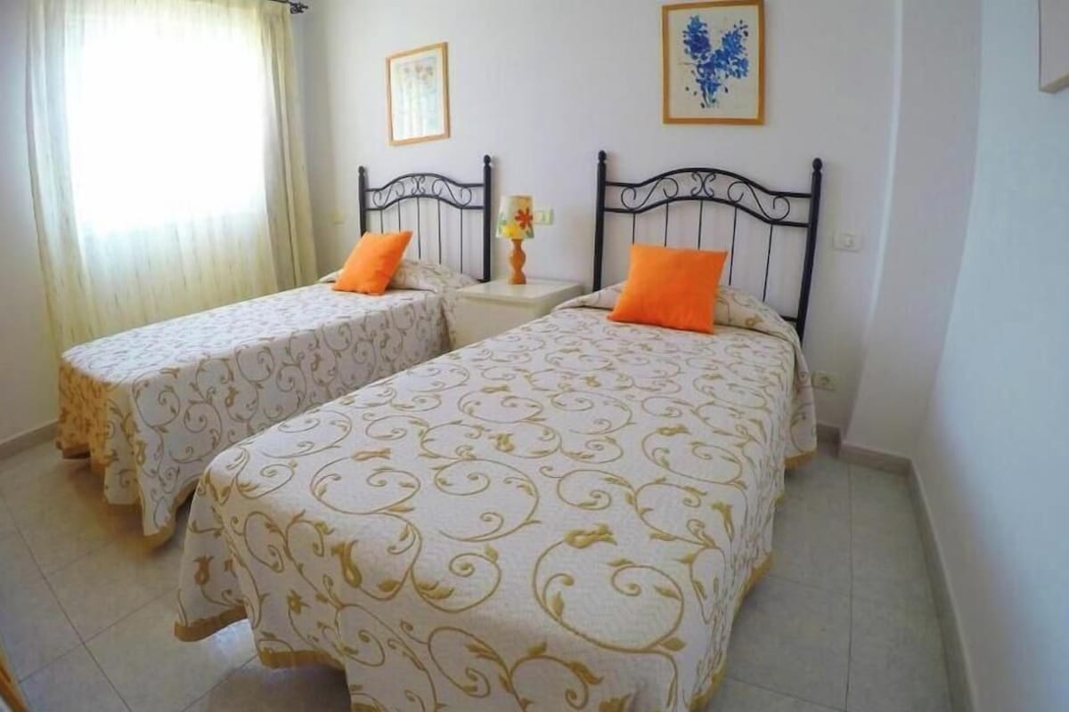 A room with two beds, each with a patterned bedspread and an orange pillow. There is a nightstand with a lamp between the beds.