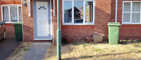 A residential brick house with a white door, two windows, and a green trash bin.
