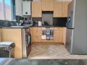 Una cocina compacta con armarios de madera, un refrigerador de acero inoxidable, un microondas, una lavadora y un piso con patrón de espiga.