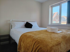 A single bed with white bedding and a brown blanket, a black pillow, and a window with a view of buildings. - 2‑Bedroom Home in Walsall – Ideal Location for Work, Study & Travel (Walsall)