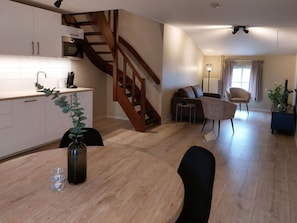 Una cocina moderna con armarios blancos, una escalera de madera, un comedor con una mesa redonda y sillas, y una sala de estar con un sofá y una lámpara.
