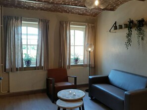 Ein Wohnzimmer mit Backstein-Decke, zwei mit Vorhängen versehenen Fenstern, einem Heizkörper, einem Sofa, einem Sessel und einem kleinen Tisch mit Kerzen.