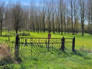 A metal gate with the inscription 'De Boerderijen' in the foreground, surrounded by a grassy field and trees in the background.