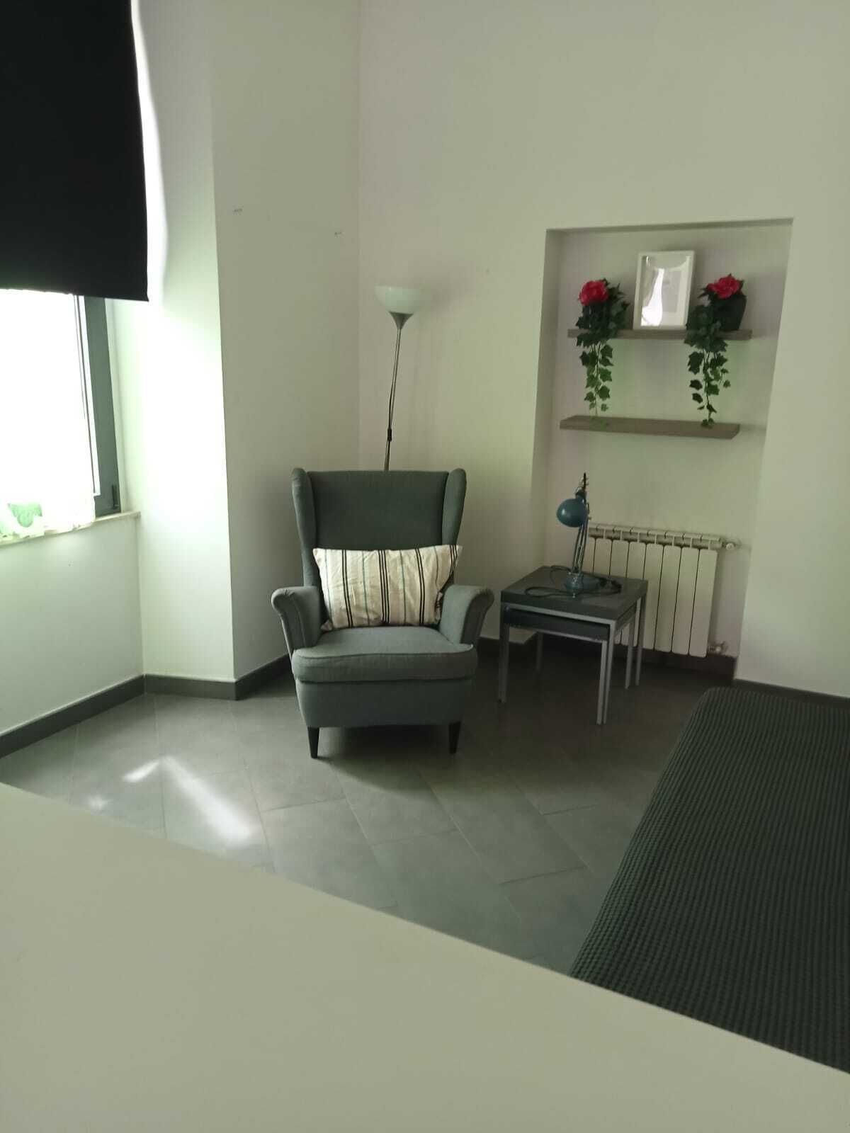 Een kamer met een grijze fauteuil, een klein tafeltje, een radiator en een raam met zonwering.