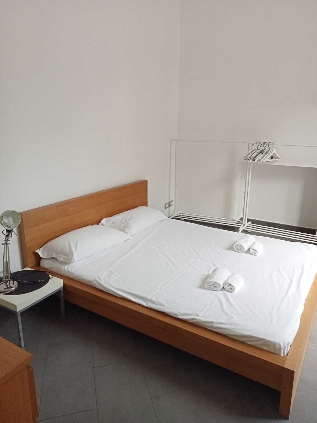 A single bed with white linens and two rolled towels, a bedside lamp, and a mirror with a towel rack.