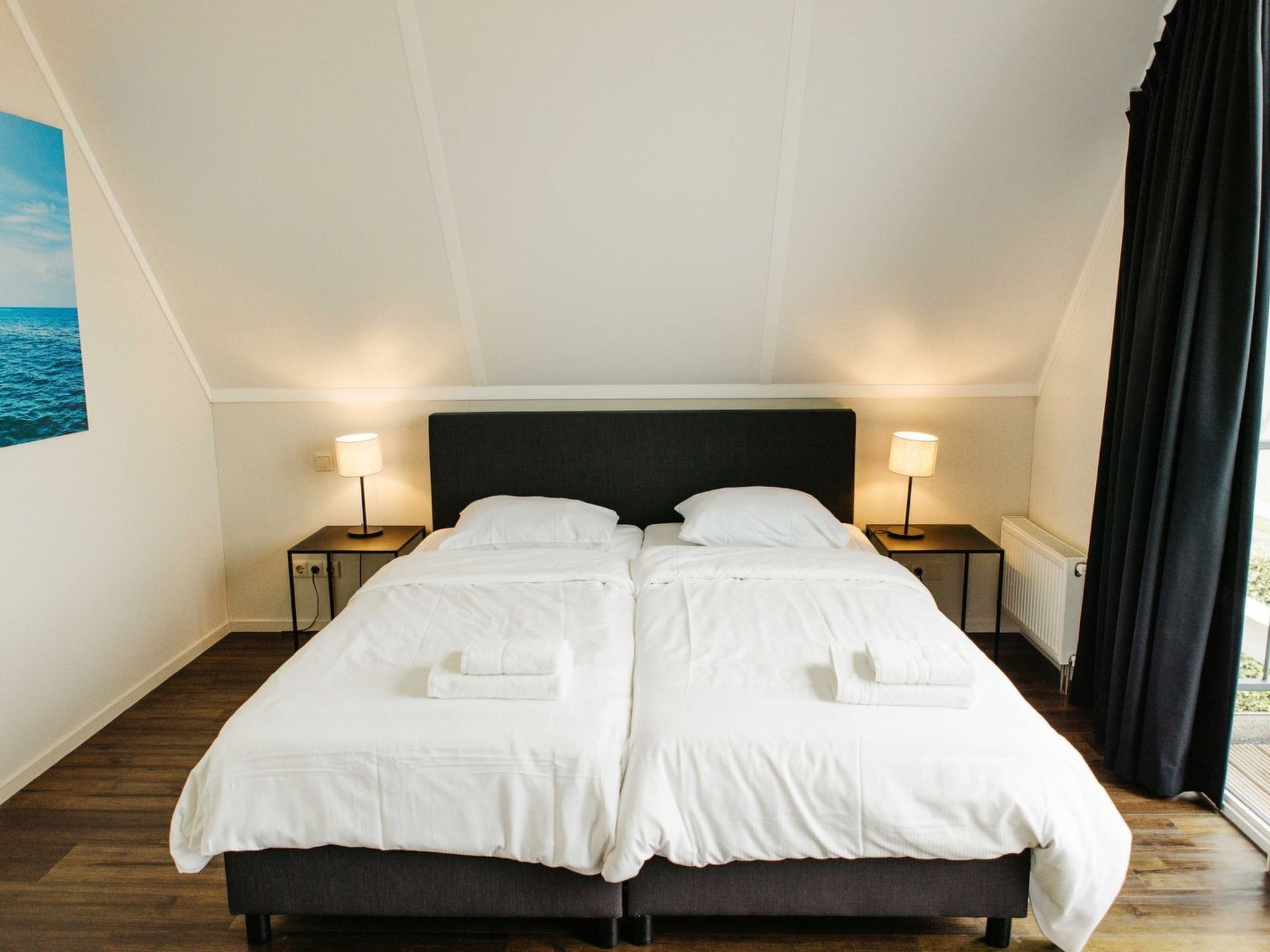 Cama doble con cabecera oscura en una habitación con suelo de madera y vista al mar por una ventana.