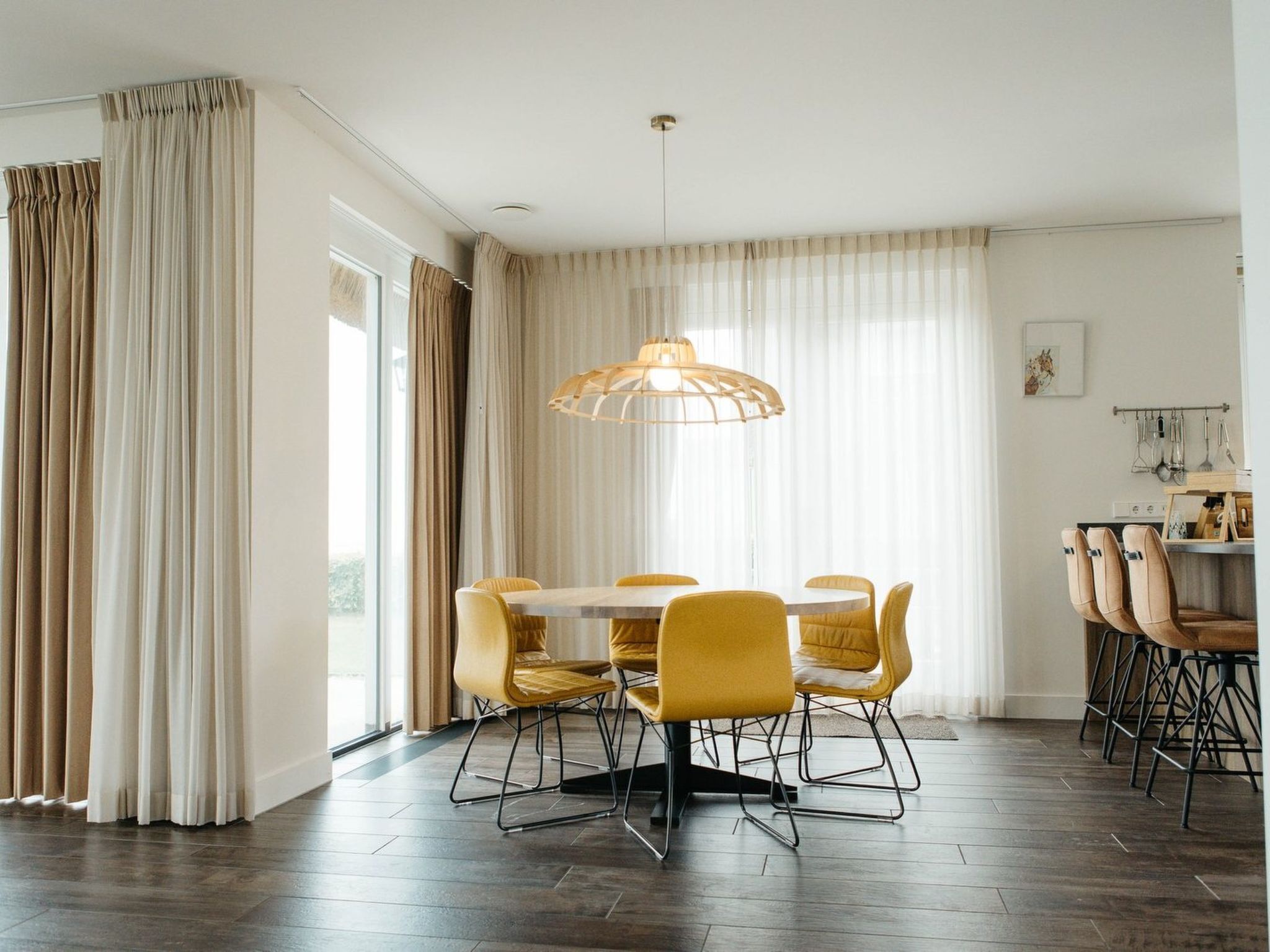 Un comedor con una mesa redonda y sillas amarillas, una lámpara colgante y vistas amplias a través de ventanas grandes.