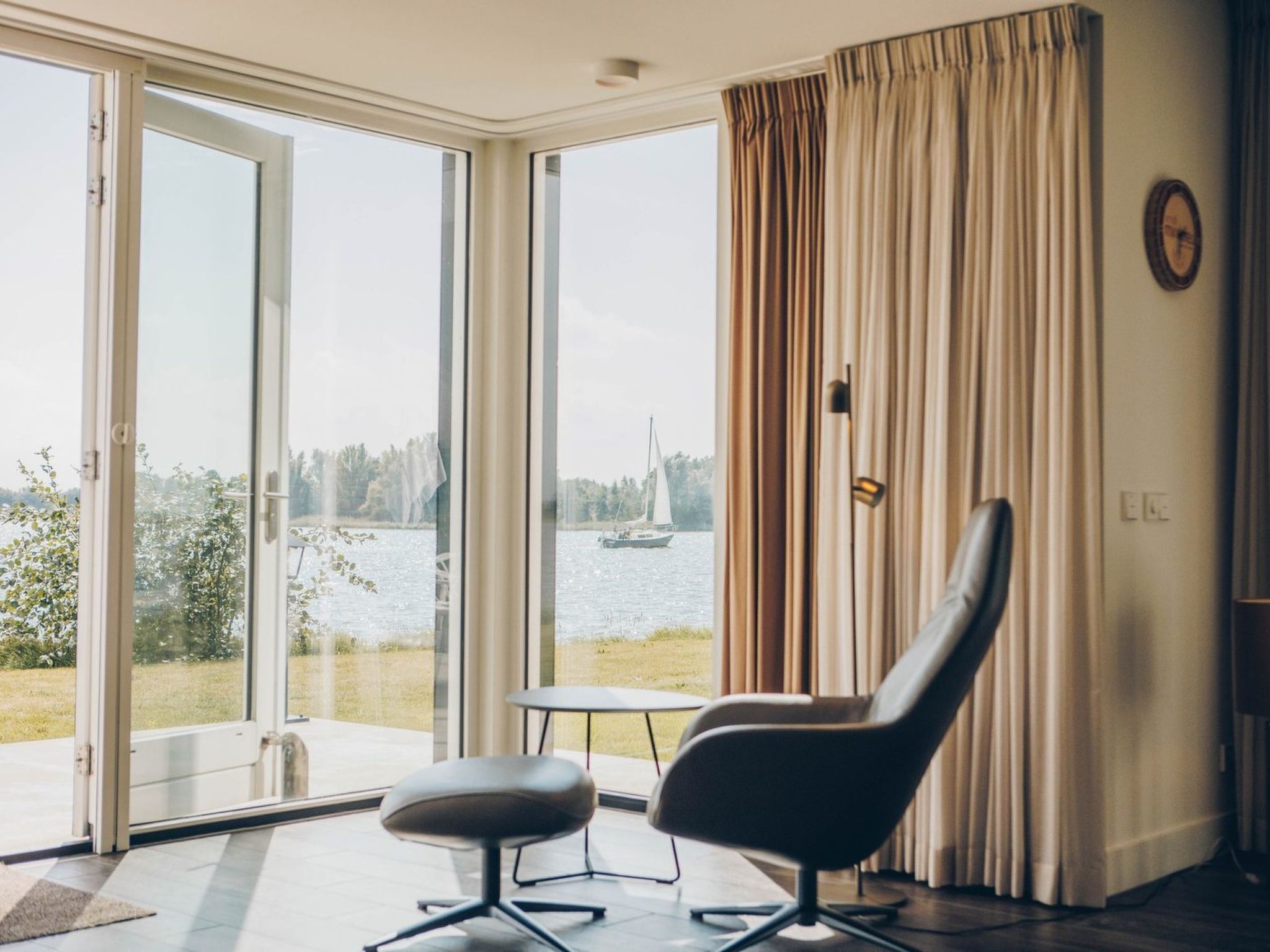 Habitación con un gran ventanal corredera que da a un balcón con vista a un lago y un velero. Hay una silla y una mesita en el balcón.