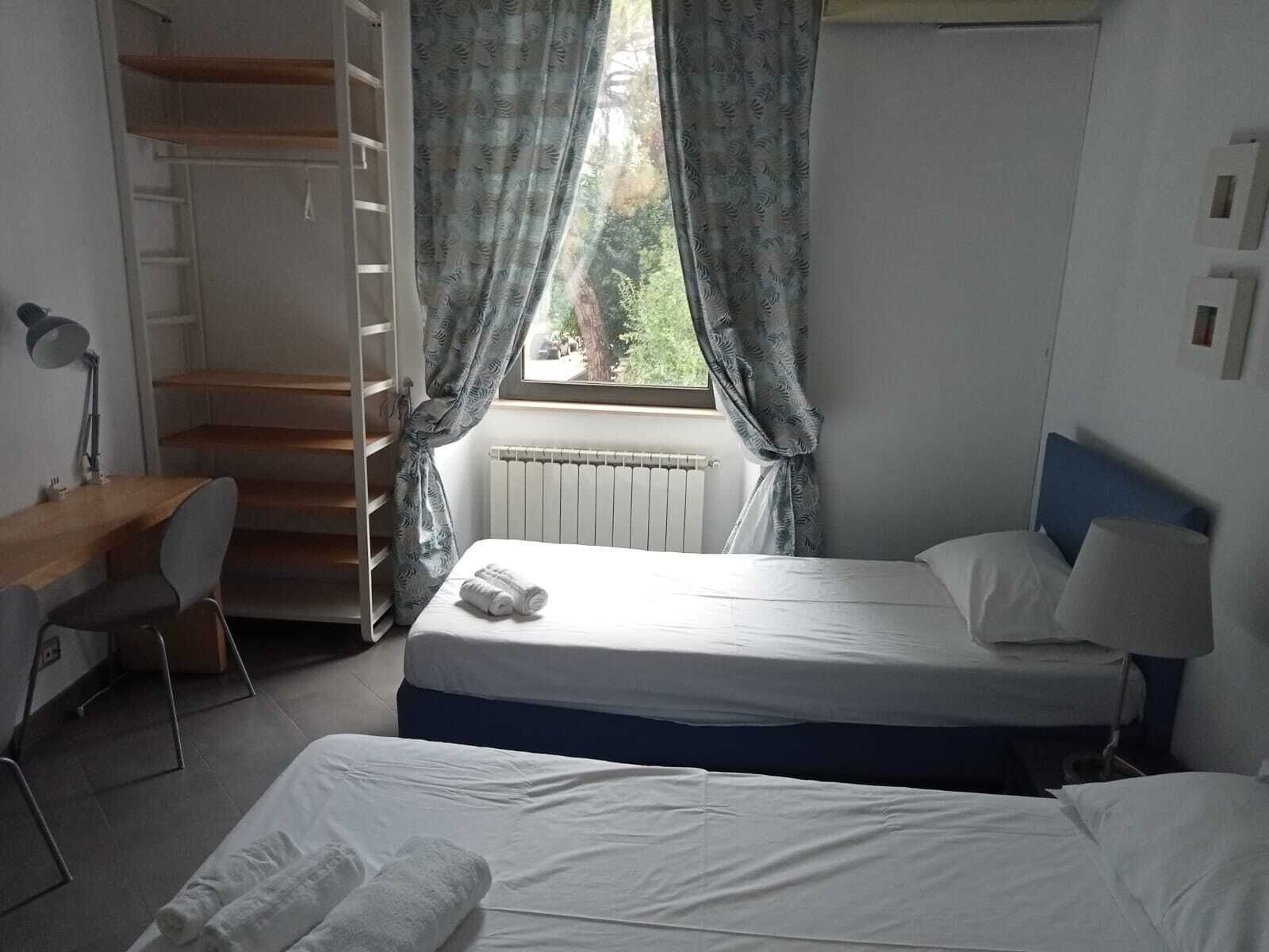 Una camera da letto con un letto, una scrivania e una sedia. Si vede una finestra con tende, un termosifone e una scala.