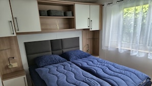 A bedroom with a bed, a nightstand, a shelf with storage boxes, and a window with curtains. - Beach Resort Kamperland - 246 by Interhome (Kamperland)