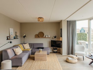 A modern living room with a grey sofa, a wooden coffee table, a flat-screen TV, and a large window with curtains.