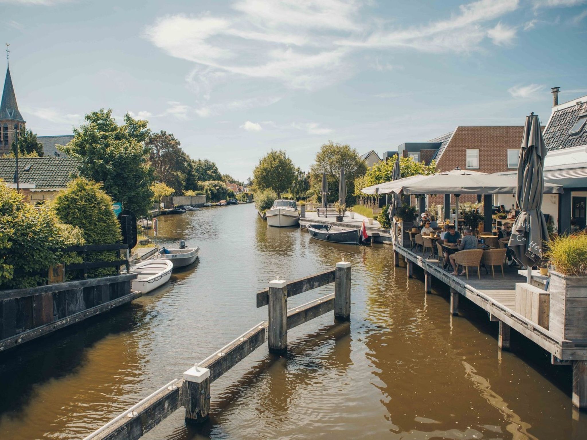 Un canal con barcos amarrados, un puente de madera y un café junto al río con terraza.