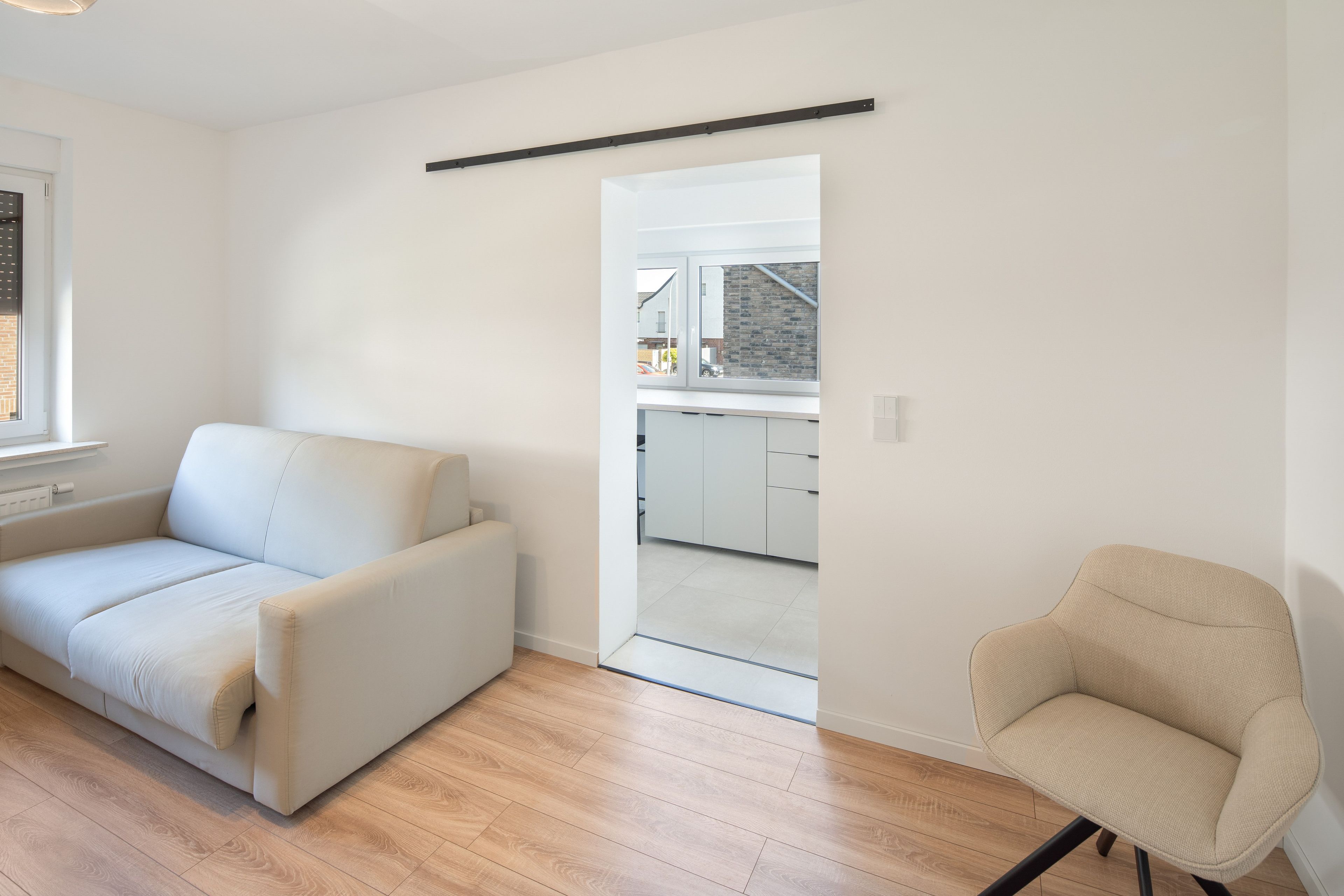 A modern living room with a sofa and a chair, a kitchen visible through an open door, and a window with a view of the outside.