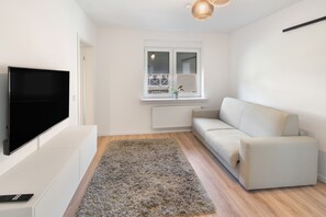 Ein modernes Wohnzimmer mit Fernseher, einem weißen Sofa, einem flauschigen Teppich in Grau und einem Fenster mit Blick auf einen Balkon.