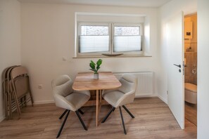 Habitació petita amb taula i dues cadires, un radiador, una finestra amb persianes i una porta que dona a una altra habitació.