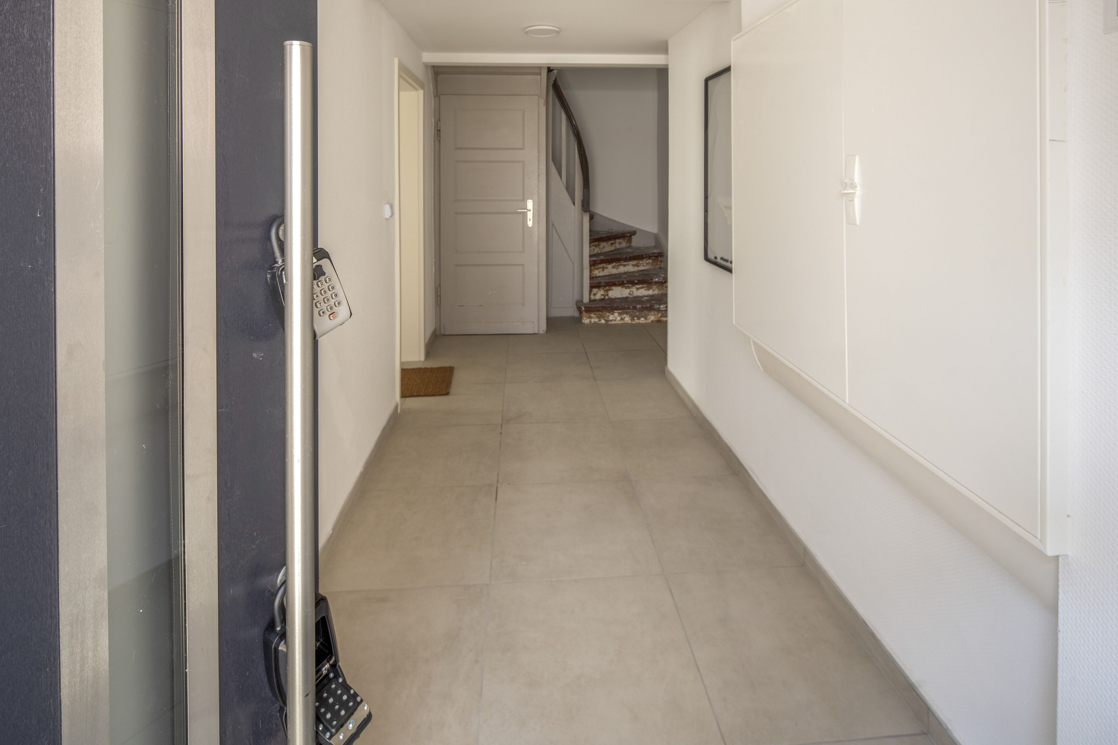 Un couloir avec une porte sur la gauche, un escalier au fond et un luminaire fixé au mur.