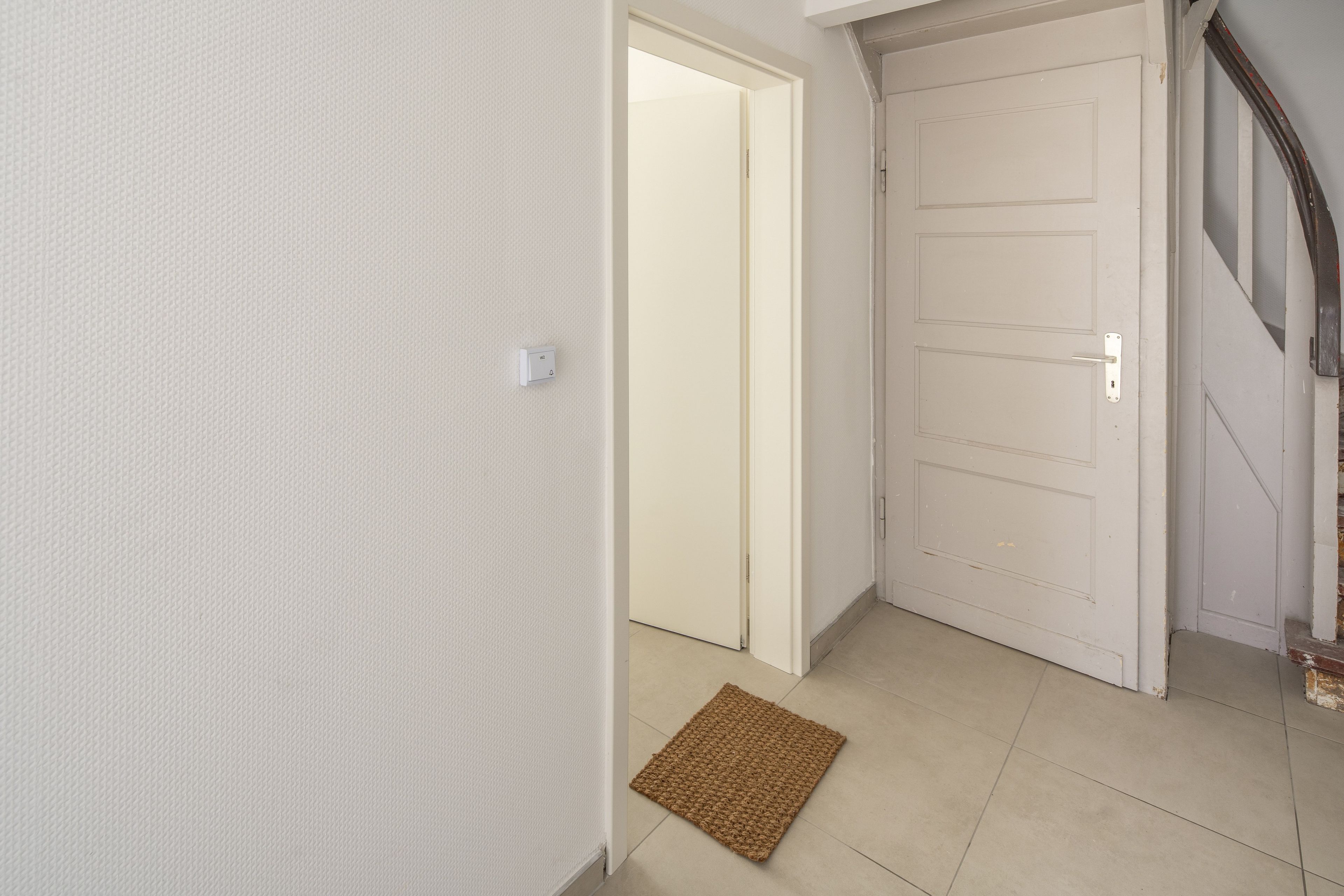 Un couloir avec un mur blanc à texture, une porte fermée et un paillasson marron sur le sol.