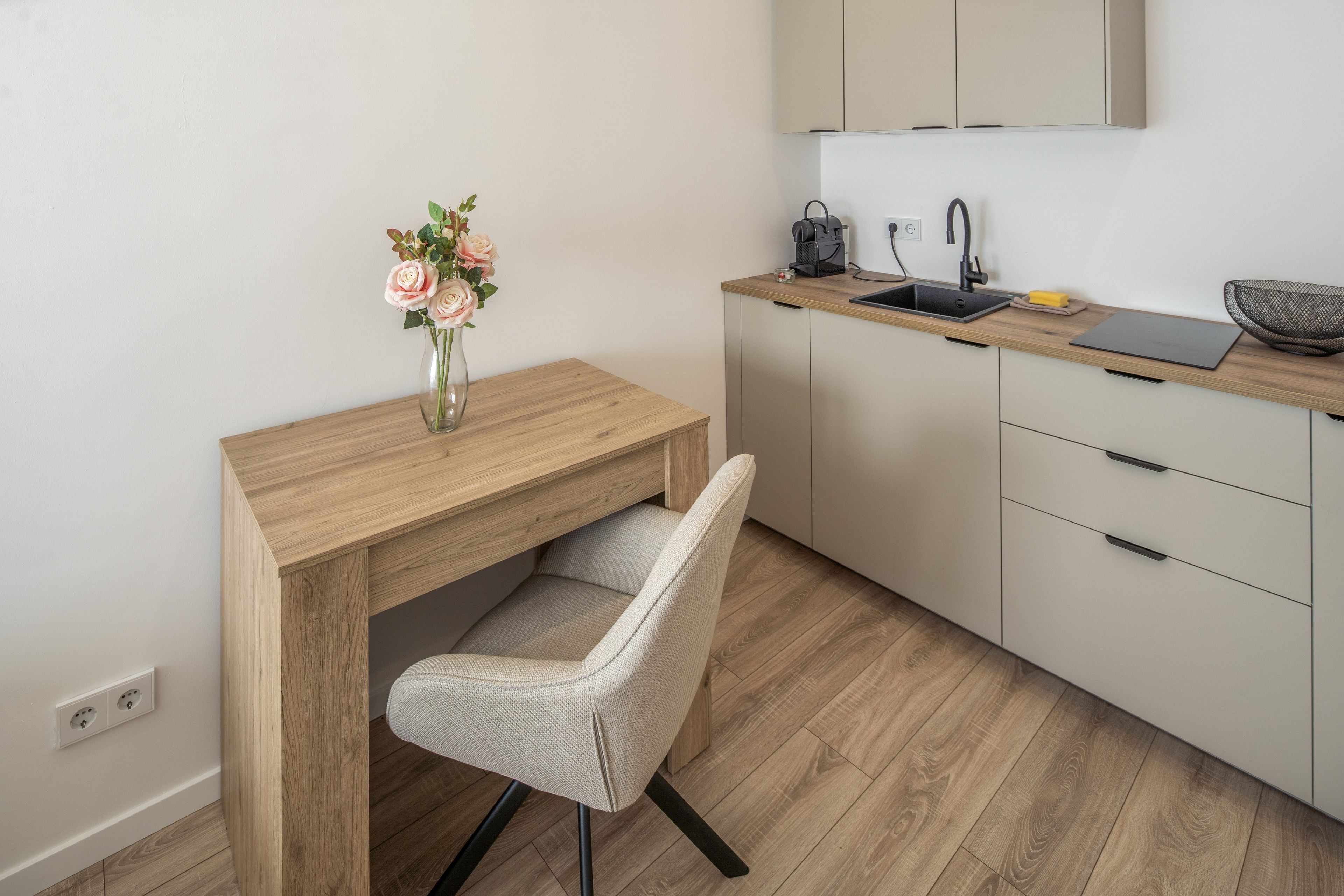 Une cuisine moderne avec un bureau en bois, une chaise, un vase avec des fleurs, une machine à café et un ordinateur portable.