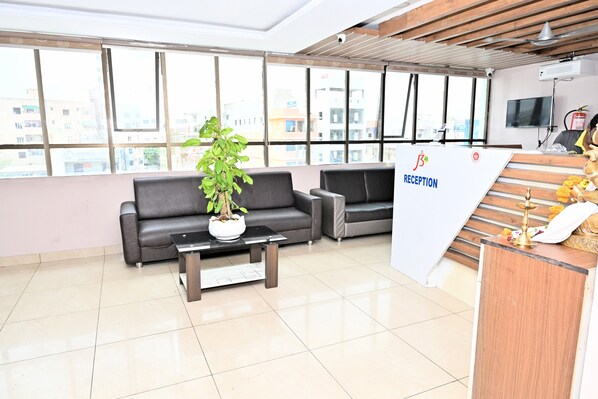 A reception area with a reception desk, a black leather sofa, a potted plant, and a view of buildings through large windows.