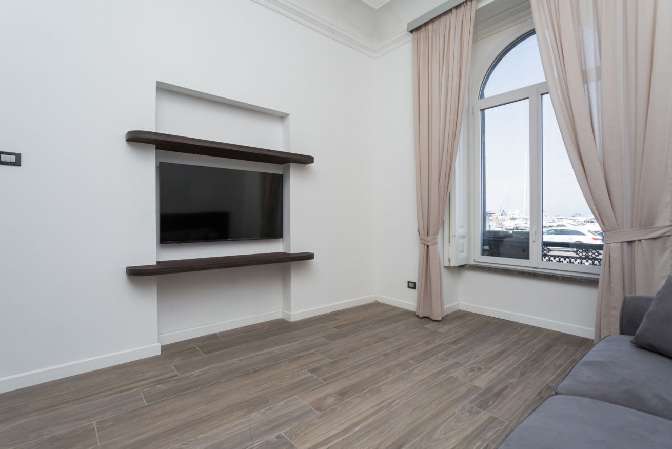 A modern living room with a flat-screen TV mounted on a wall, a window with curtains, and a grey sofa.