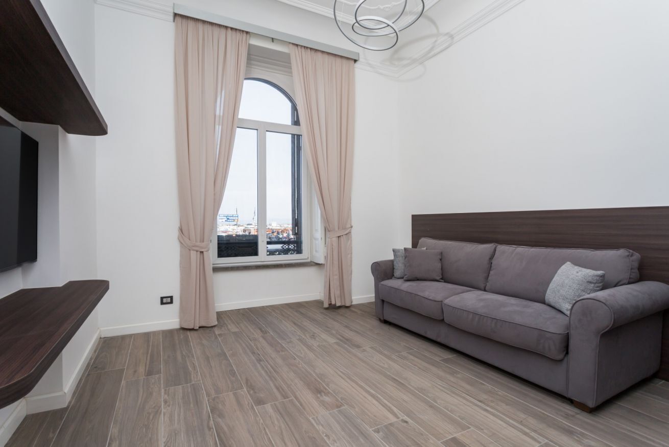 Ein modernes Wohnzimmer mit grauem Sofa, einem Fenster mit Vorhängen und einem an der Wand befestigten Flachbildfernseher.
