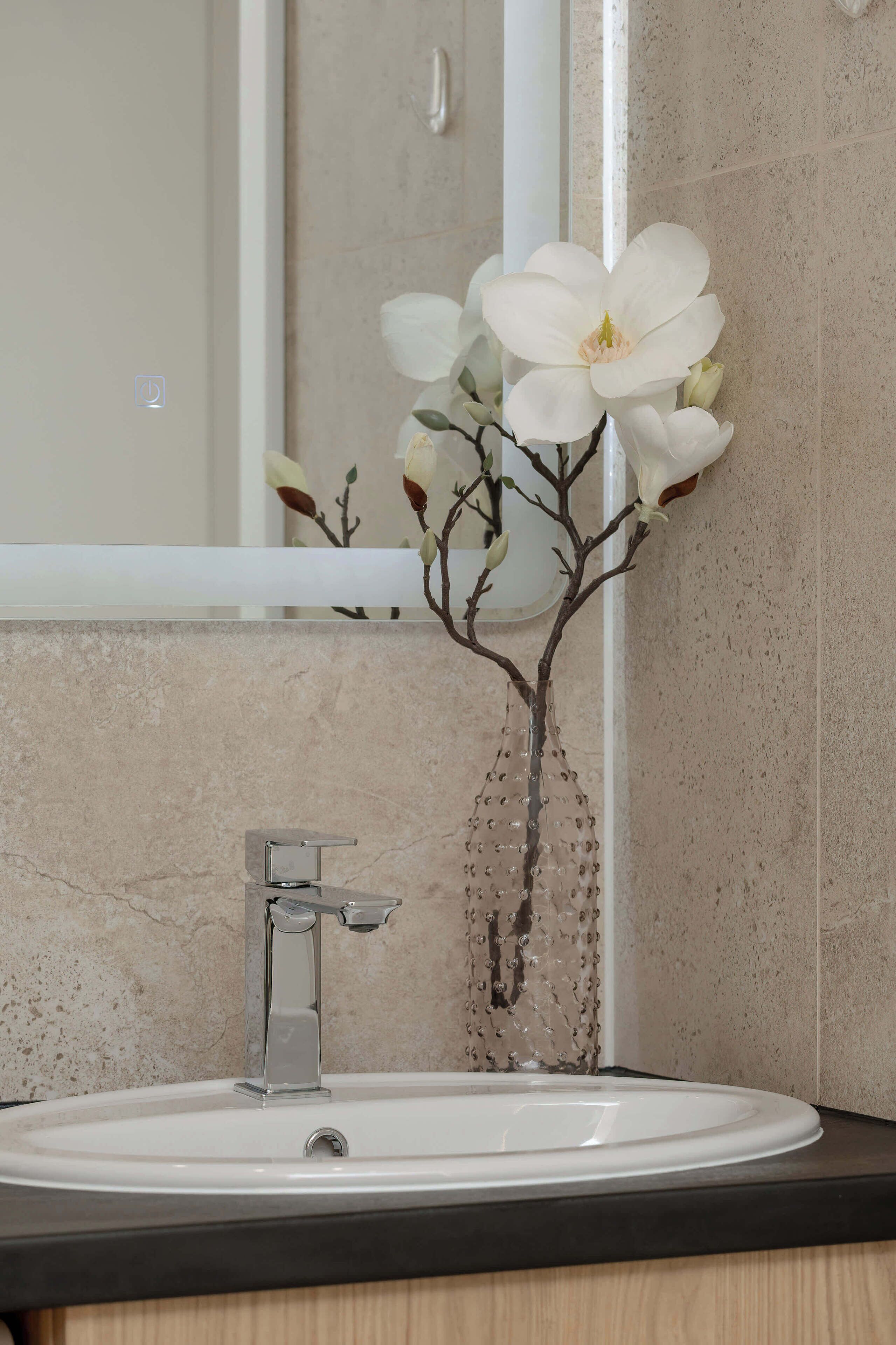 Un lavamanos de baño con una grifería moderna, un espejo encima y una rama decorativa con flores blancas.