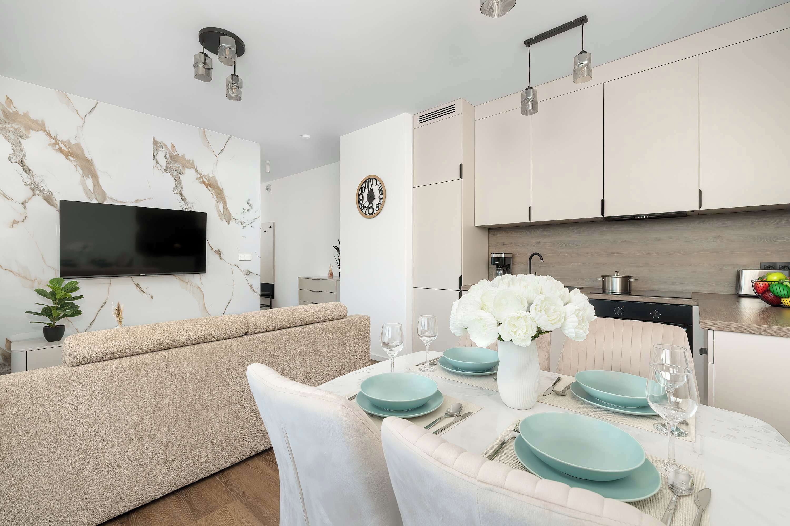A modern kitchen with white cabinets, a flat-screen TV mounted on the wall, and a dining area with a table set for two.