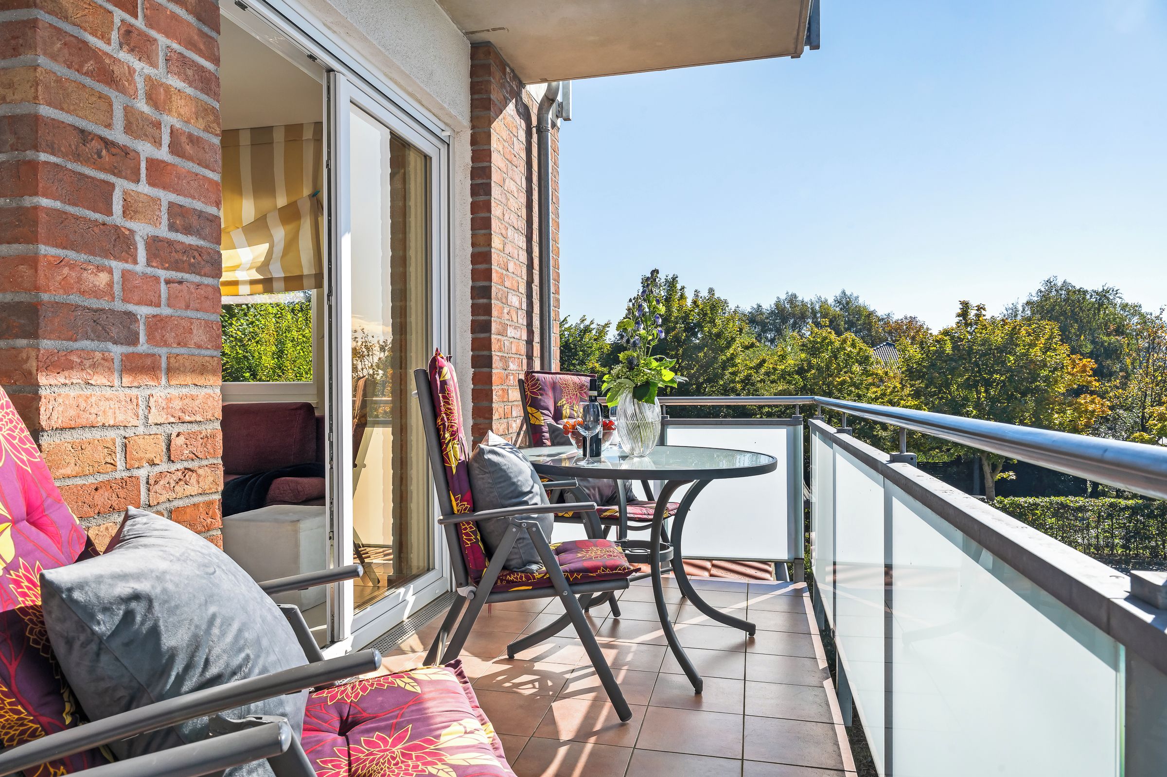 Een balkon met een tafel en stoelen, een bank en uitzicht op bomen en gebouwen.