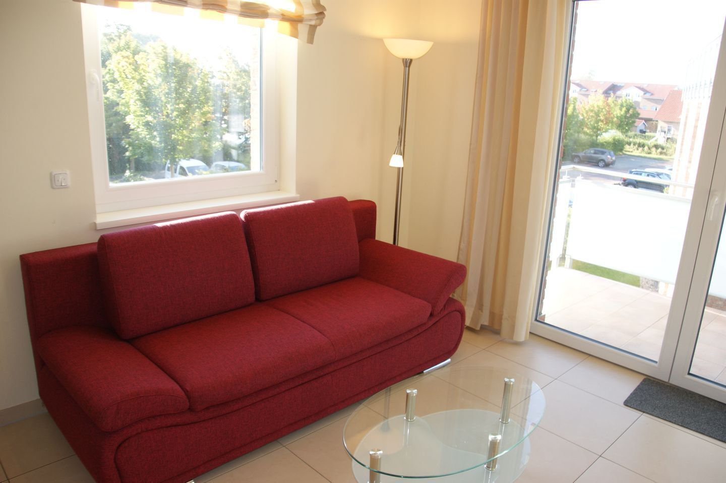 A red sofa in a room with a glass coffee table, a floor lamp, and a view of a parking area through a window.