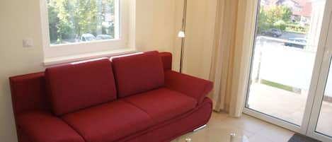 A red sofa in a room with a glass coffee table, a floor lamp, and a view of a parking area through a window.