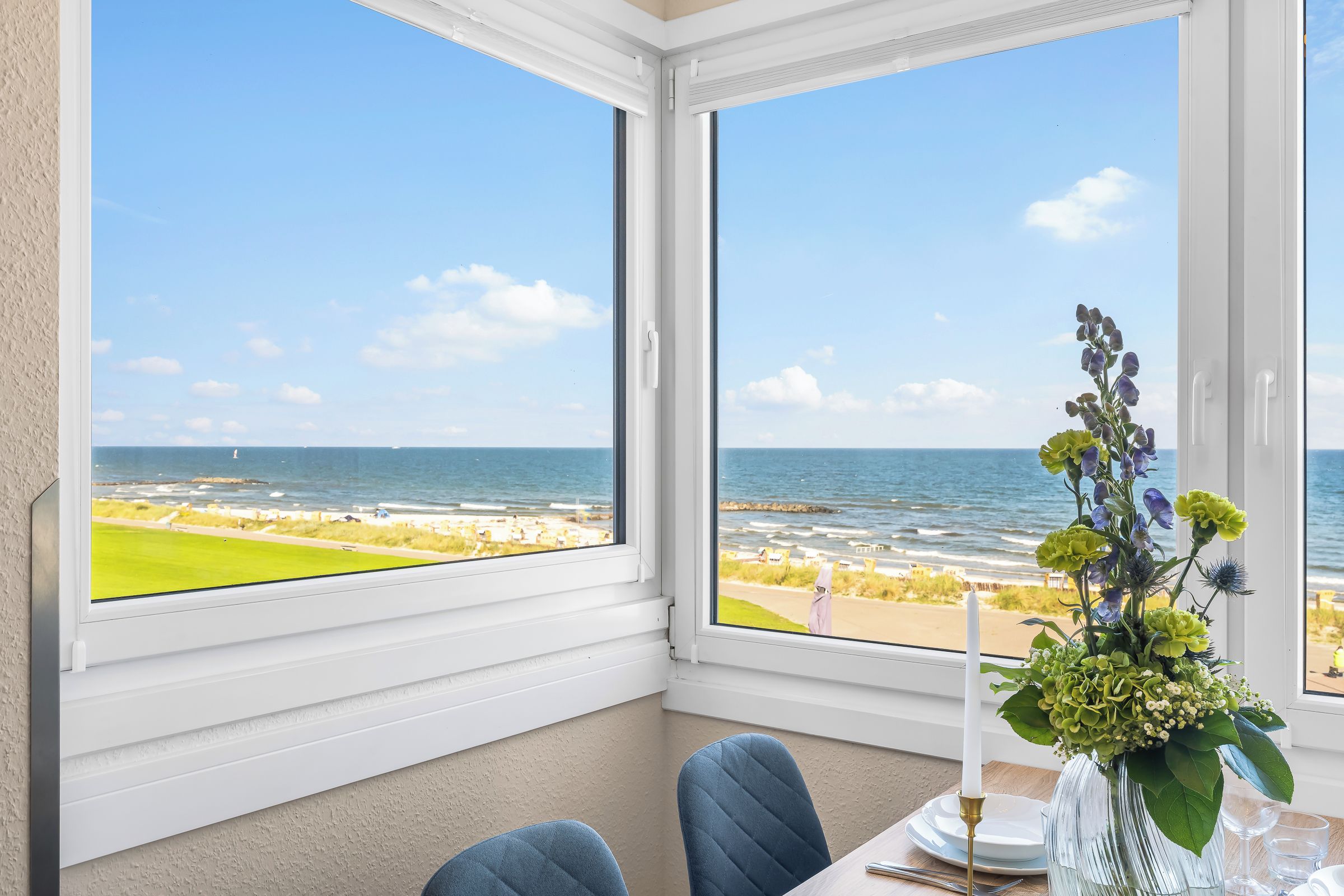 A room with a large window overlooking a beach and ocean, a table with a vase of flowers, and a chair.