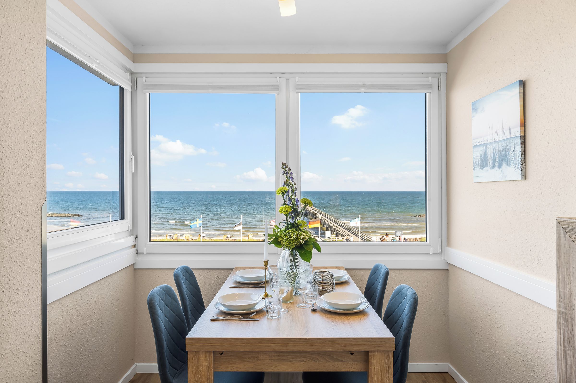 Un coin repas avec une table dressée pour deux personnes, donnant sur une plage où flottent des drapeaux et où se trouve une jetée.