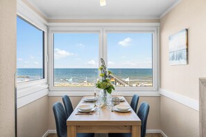 A dining area with a table set for two, overlooking a beach with flags and a pier. - Holiday Apartment Strandblick – Sea Views, Relaxation & Holiday Bliss (Schönberg (Holstein))