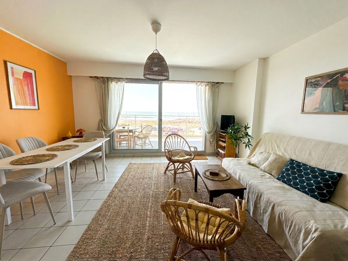 A living room with a sofa, dining table, chairs, and a view of the beach through the window.