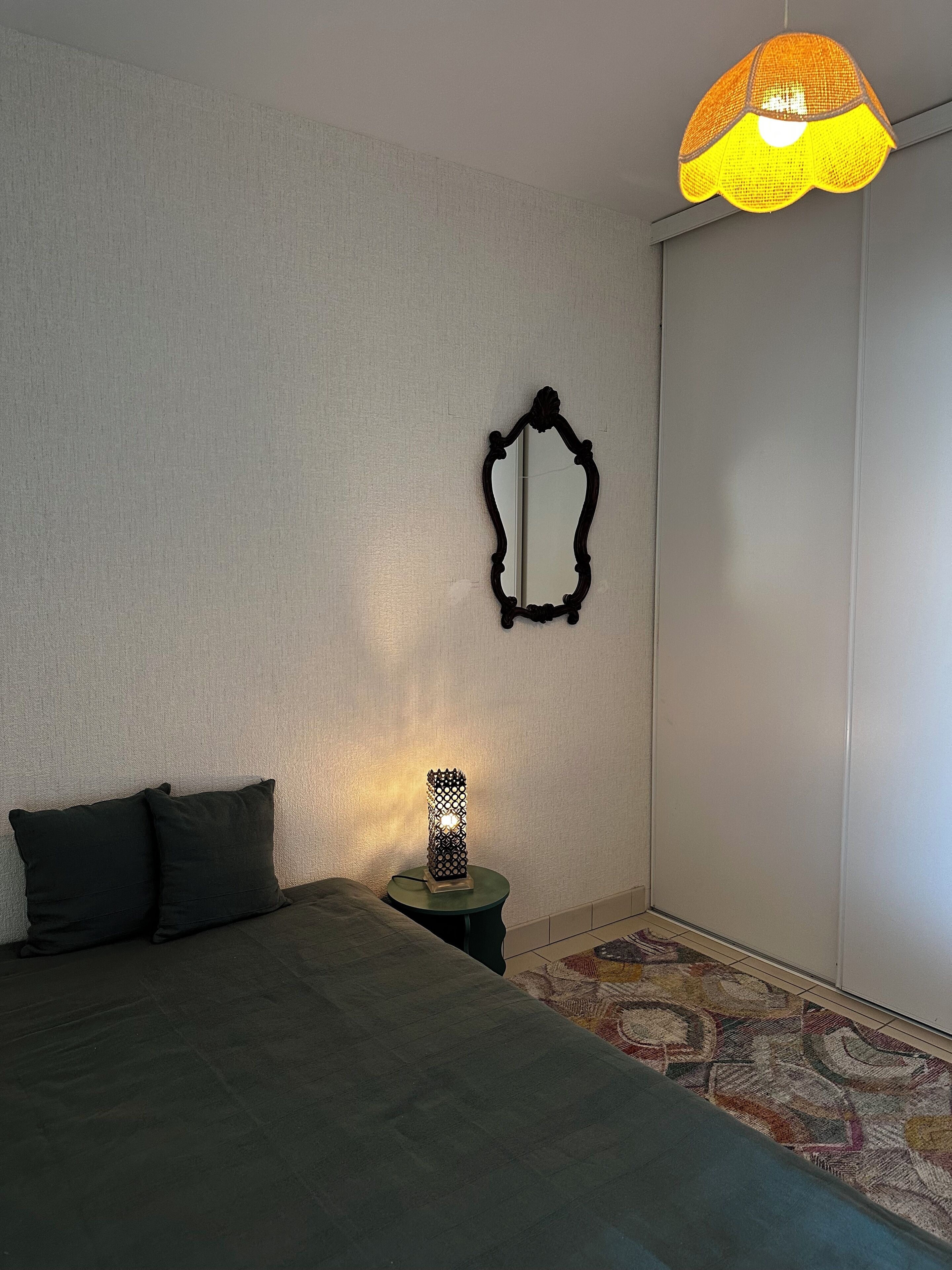Een slaapkamer met een bed, een klein tafeltje met een lamp, een spiegel aan de muur en een kamerkleed met een patroon.