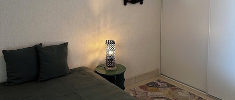 Ein Schlafzimmer mit einem Bett, einem kleinen Tisch mit Lampe, einem Spiegel an der Wand und einem gemusterten Teppich.