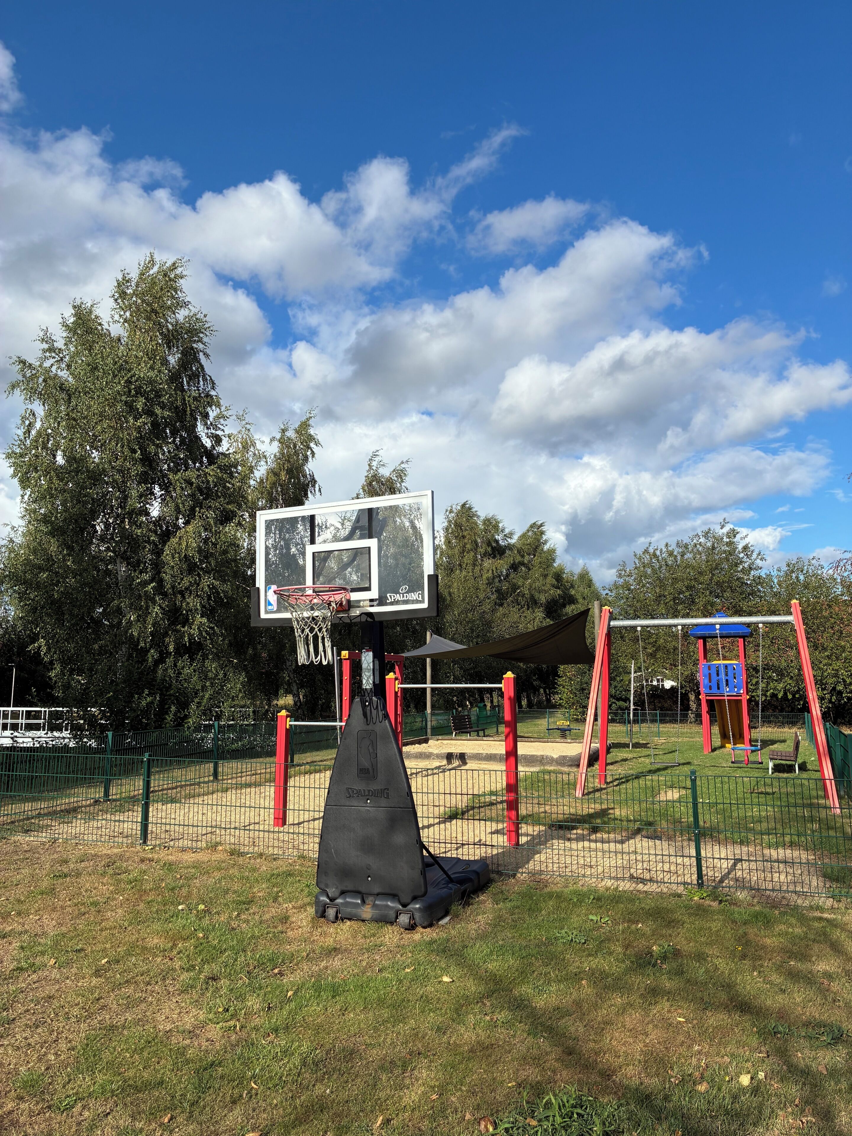 A basketball court with a Spalding hoop, a swing set, and a fenced playground area.
