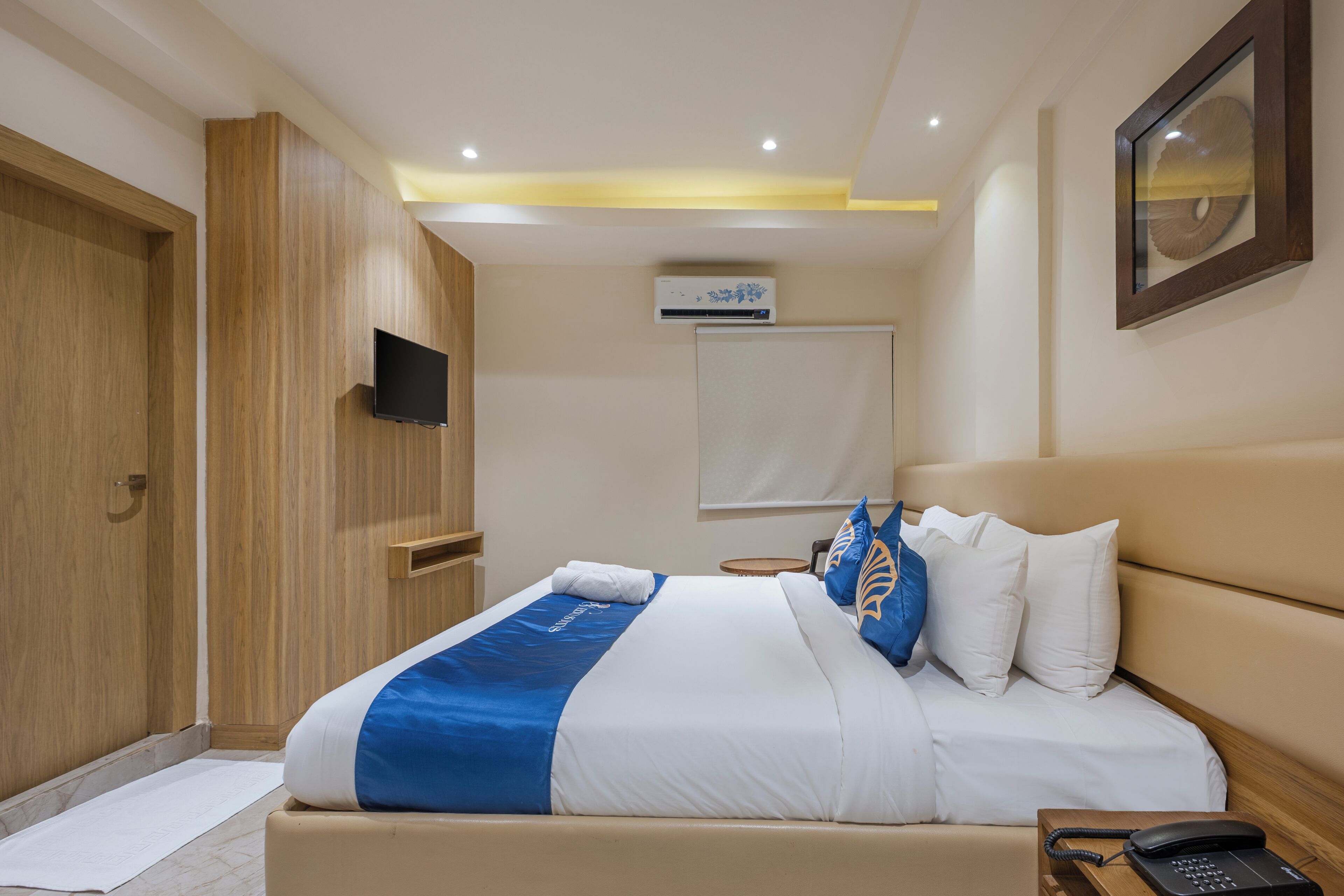 Una habitación de hotel con cama, mesita de noche, teléfono, aire acondicionado y televisor montado en la pared.
