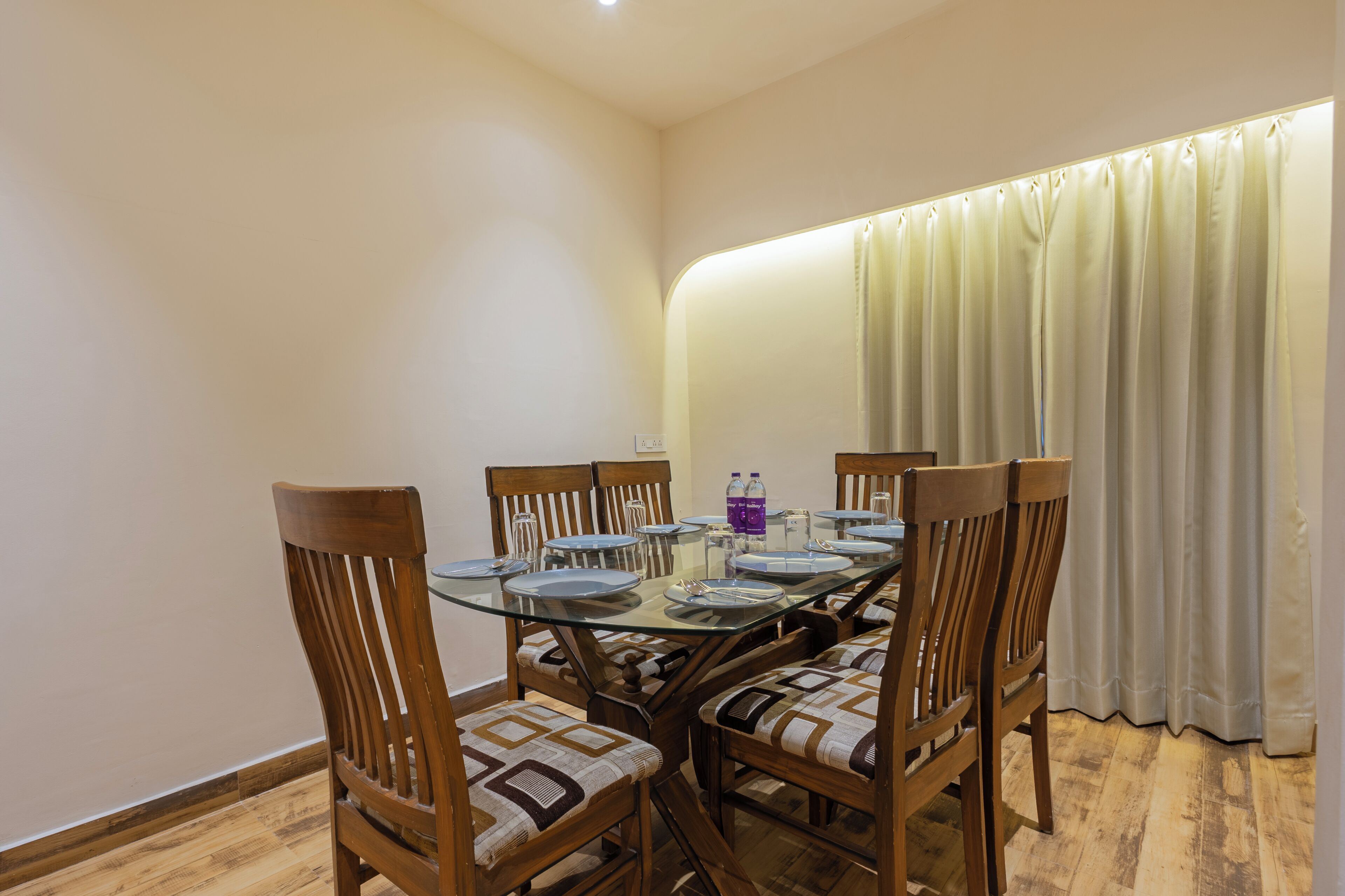A dining area with a glass table, wooden chairs with patterned cushions, and a curtain on the right.
