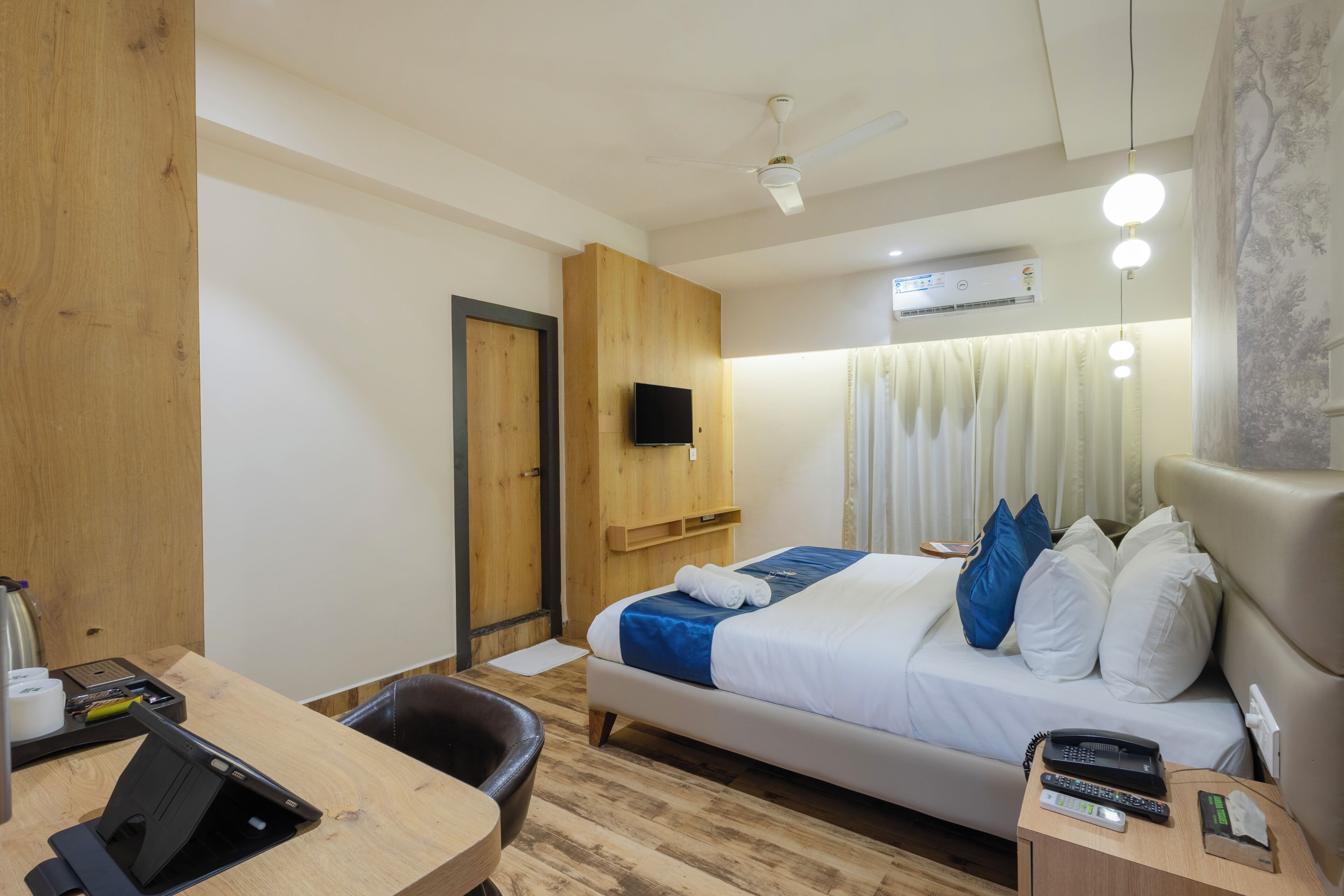 A hotel room with a bed, a desk with a laptop, a chair, a TV, and an air conditioning unit.